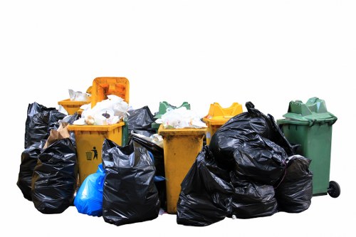 A collection of eight wheelie bins and rubbish bags positioned outdoors on a paved surface, with the bins in various colours including yellow, green, and black. The yellow bins contain mixed waste, with some overflowing, and are built from plastic with textured surfaces. The green bin, also made of plastic, appears to be a general waste receptacle. Several black rubbish bags, filled with waste, are piled in front and around the bins, some tied at the top and others partially open. The waste visible includes white paper and packaging materials, suggesting general household or office clearance rubbish. The scene is set on a paved area with no background context, possibly a driveway or loading zone typical for rubbish collection in Northolt, UK. The lighting is neutral, providing clear visibility of the textures, colors, and materials of the bins and rubbish bags, consistent with realistic outdoor environment conditions. This image visually relates to waste collection and rubbish disposal services offered by companies like Office Clearance Northolt in the local area.