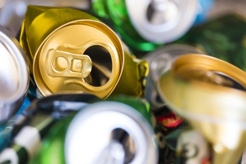 Close-up view of a pile of crushed aluminum cans in various colors, including gold, silver, and green, with some cans featuring visible pull-tabs and smooth metallic surfaces. The cans are piled together on a surface, with some overlapping each other, and are partially out of focus towards the edges of the image. The environment suggests an outdoor setting, potentially on a pavement or driveway in Northolt, with natural lighting illuminating the metallic textures. This image captures the type of waste material that a rubbish removal service like Office Clearance Northolt might handle during a local clearance or recycling collection, emphasizing the presence of discarded beverage cans typically collected in civic or commercial waste pickups in the area.