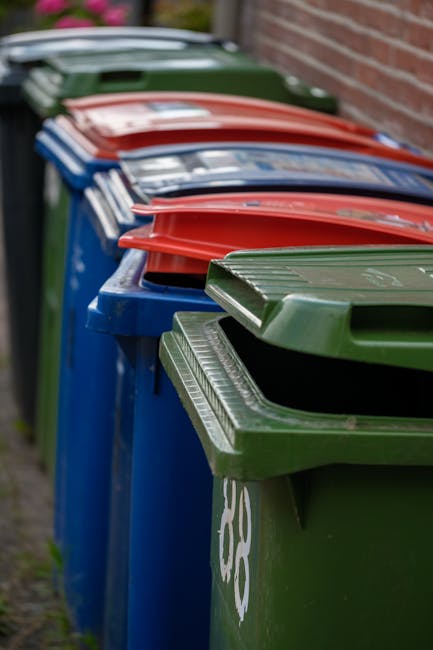 A green, large wheeled rubbish bin filled with red and black bags of garden waste, positioned on a concrete pavement next to a dark blue brick wall. The bags are labeled with gardening product informa