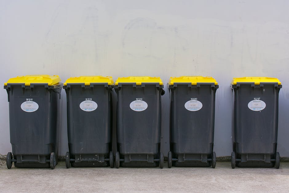 A row of five large wheeled rubbish bins with dark grey bodies and bright yellow lids positioned against a plain, light-colored wall. Each bin features a white oval label with black text and identific