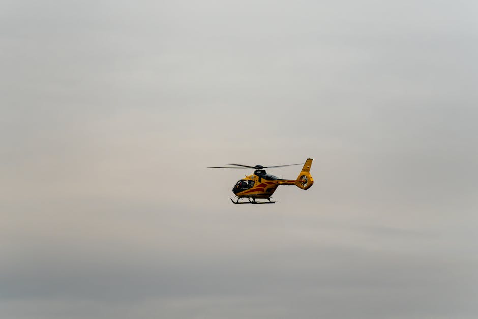 A yellow and black helicopter with a streamlined fuselage and rotor blades is flying against a cloudy gray sky. The helicopter's tail boom extends to a vertical stabilizer with a small tail rotor. Its