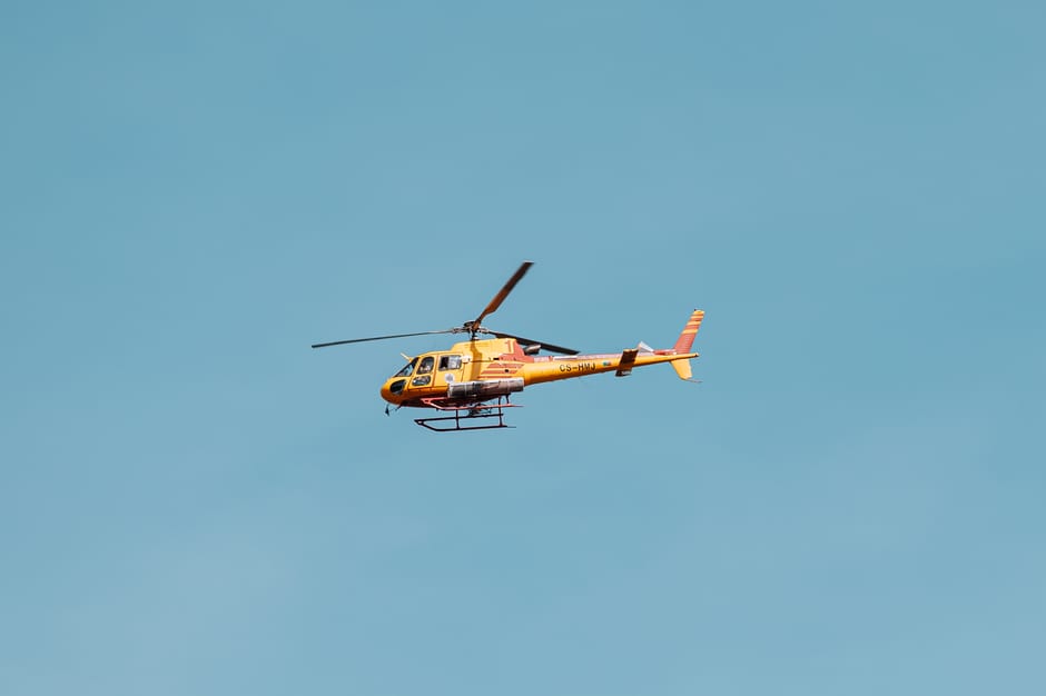 A yellow and black helicopter with a streamlined fuselage and rotor blades is flying against a cloudy gray sky. The helicopter's tail boom extends to a vertical stabilizer with a small tail rotor. Its