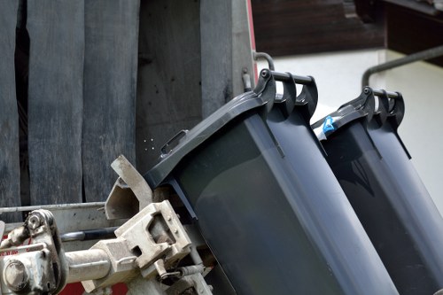 Image showing two dark grey wheelie bins attached to a metal bin lorry, prepared for rubbish collection. The bins are made of durable plastic with slightly textured surfaces, and their lids are closed. The lorry's metallic mechanical arms are visible on the left side, gripping the bins securely, with some signs of wear and slight rust. The background includes a wooden fence and part of a building's exterior, suggesting an urban or suburban setting. The scene is illuminated with natural daylight, highlighting the metallic and plastic textures. This arrangement reflects a typical rubbish removal process in Northolt, with the bins positioned on a paved driveway or street frontage ready for collection by Office Clearance Northolt as part of their waste management services within the local area.