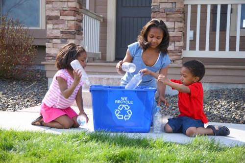 A woman and two children, a girl and a boy, are sitting on a concrete pathway in front of a residential property, participating in a rubbish collection activity. The woman, wearing a light blue sleeveless top, is pouring plastic bottles into a blue recycling bin with a 'We Recycle' label and a recycling symbol. The girl, dressed in a pink and white striped shirt with a pink skirt, is holding a plastic bottle close to her mouth, appearing to be drinking or preparing to place it into the bin. The boy, in a red T-shirt and dark shorts, is holding a plastic bottle, ready to dispose of it, and is sitting on the ground next to the woman. Behind them, there is a modern house with a grey door and a white railing on the porch, with a brick and stone facade. The surrounding outdoor environment includes a well-maintained lawn with lush green grass, small shrubs on the left, and scattered rocks along the edging of the pathway. The scene is captured in natural daylight, emphasizing the orderly and eco-conscious activity involved in rubbish and recycling handling, potentially related to office clearance services in Northolt.